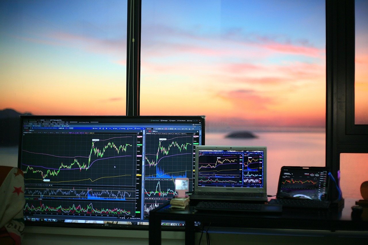 夕焼けの海を背景に複数のモニターに株価チャートを表示した室内デスク