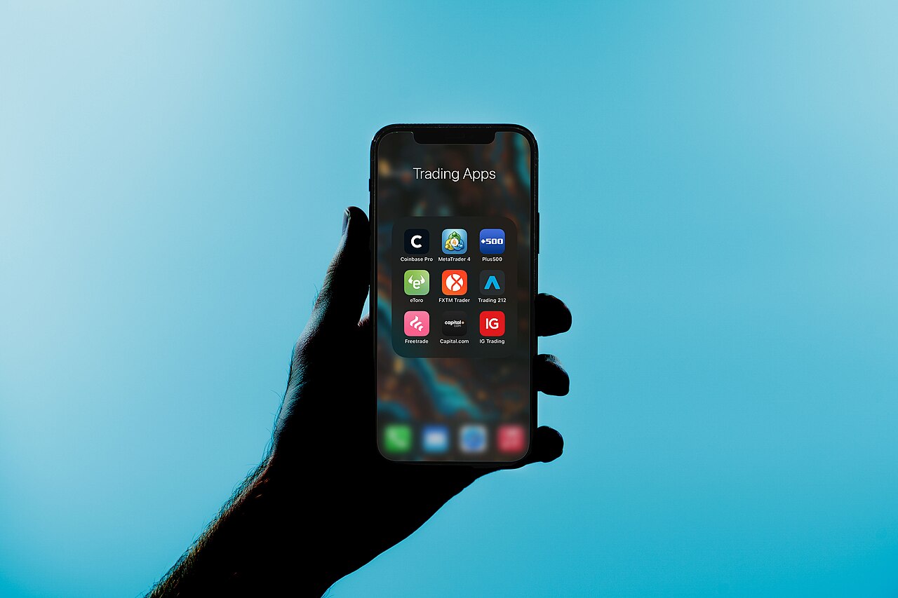 逆光のシルエットになった手がスマートフォンを持ち、画面には取引アプリのフォルダが開かれて9つのアプリアイコンが3×3で並んでいる