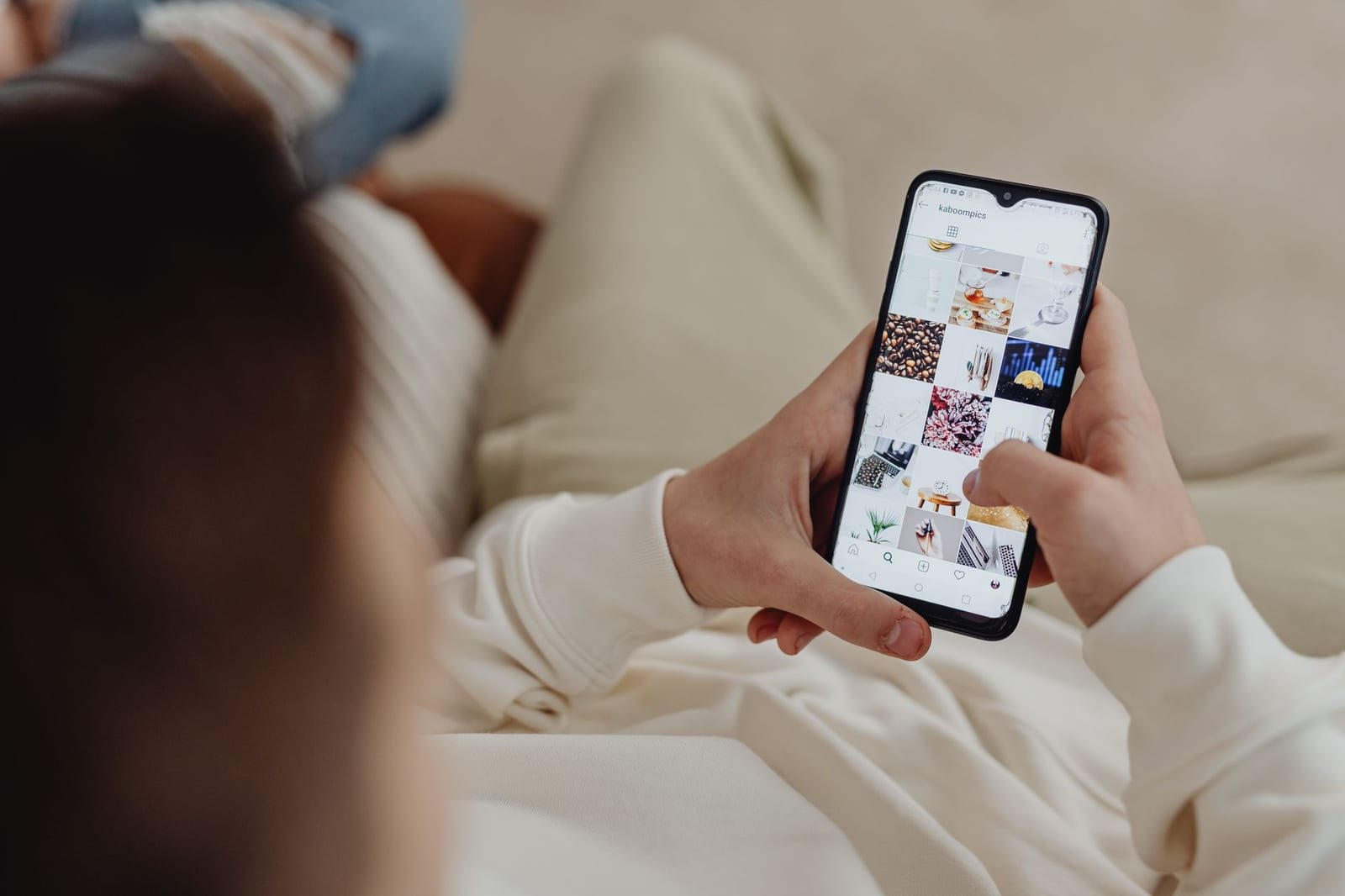 ソファに座る人物が肩越しにSNSの写真一覧が表示されたスマートフォンを操作している