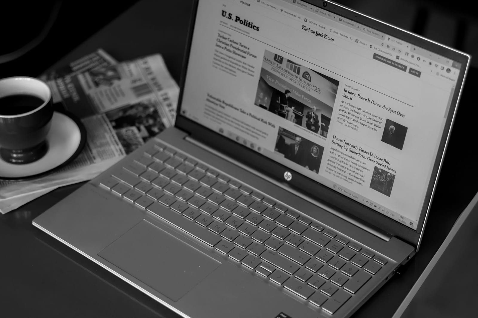コーヒーと新聞の横で開いたノートパソコンにニュースサイトが表示された室内の白黒写真。
