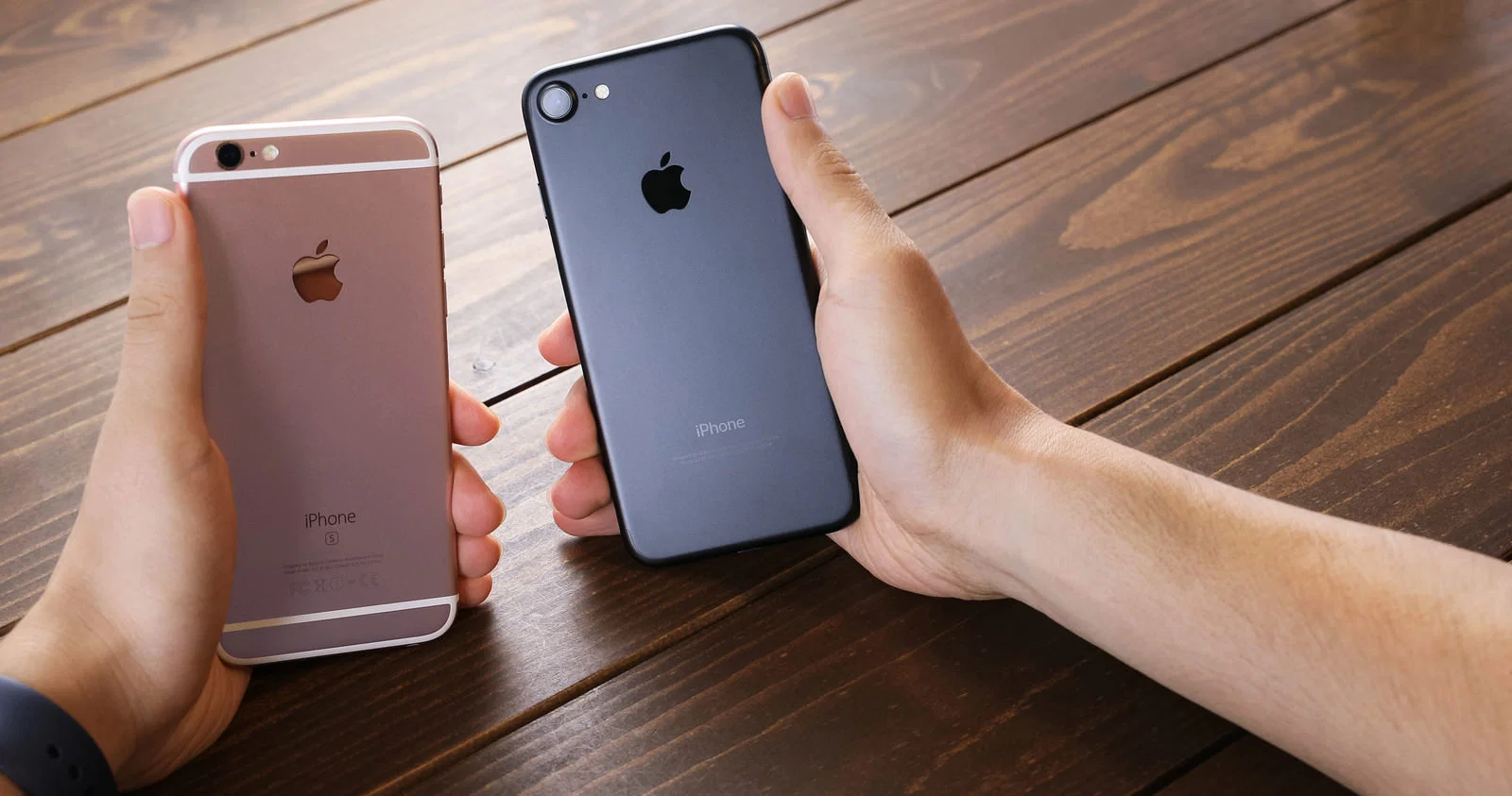 屋内の木製テーブルで両手にiPhoneを1台ずつ持ち背面を見比べている写真