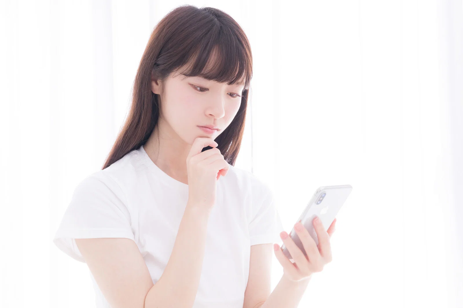 白いカーテンを背に白Tシャツの女性1人がスマートフォンを見て考え込む写真