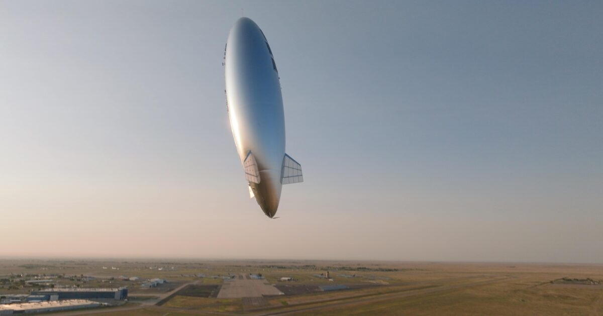 飛行場の上空に浮かぶ銀色の飛行船型機体を屋外で見上げた写真