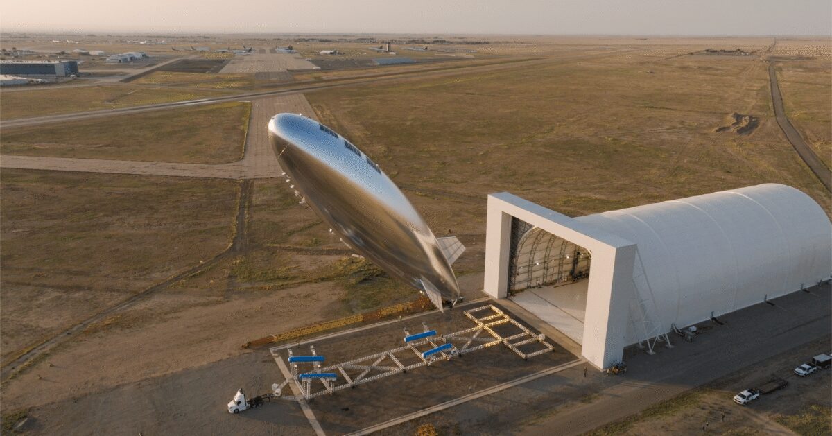 白い大型格納庫の開いた扉の前で、銀色の飛行船型の機体が機首を斜め上に向けて浮かんでいる空撮写真。手前には金属トラス状の地上設備と作業車両が並び、背景に滑走路と平原が広がる