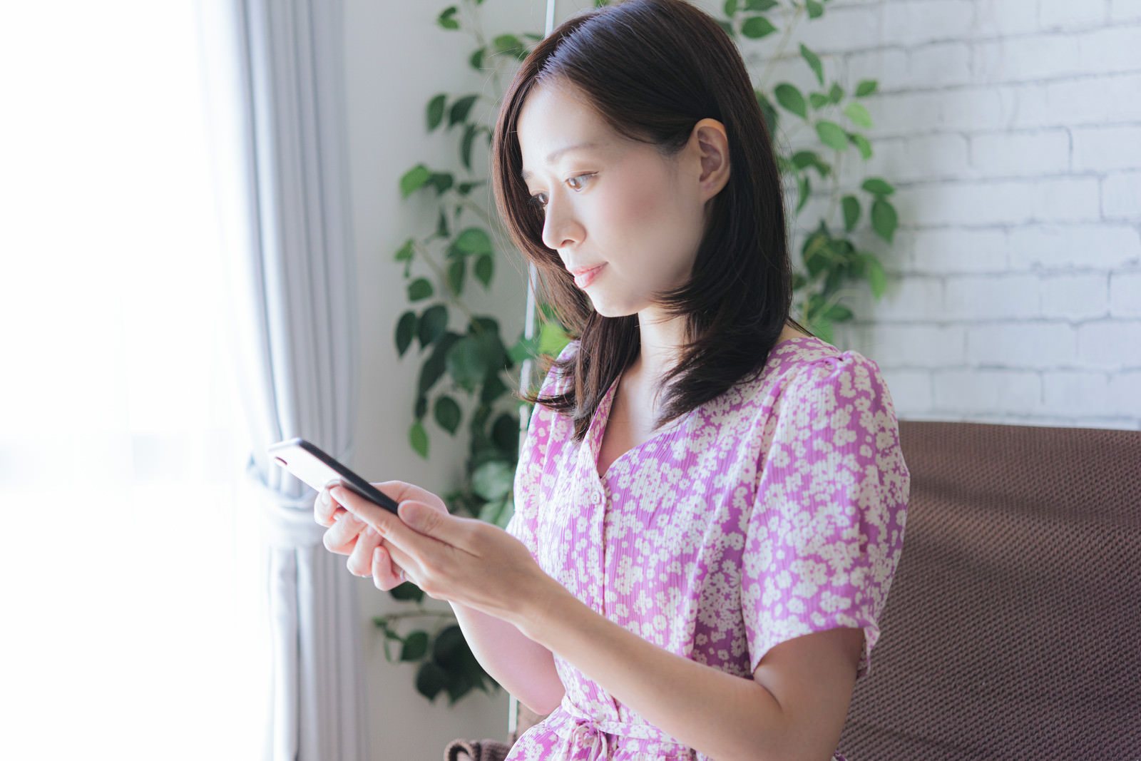 室内で女性一人がソファに座りスマートフォンの画面を見つめる