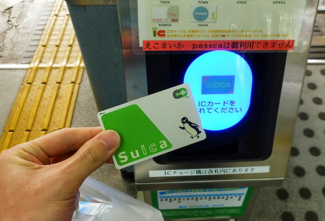 屋外の日本の駅で、手にSuicaカードをかざし改札機に読み取らせている写真