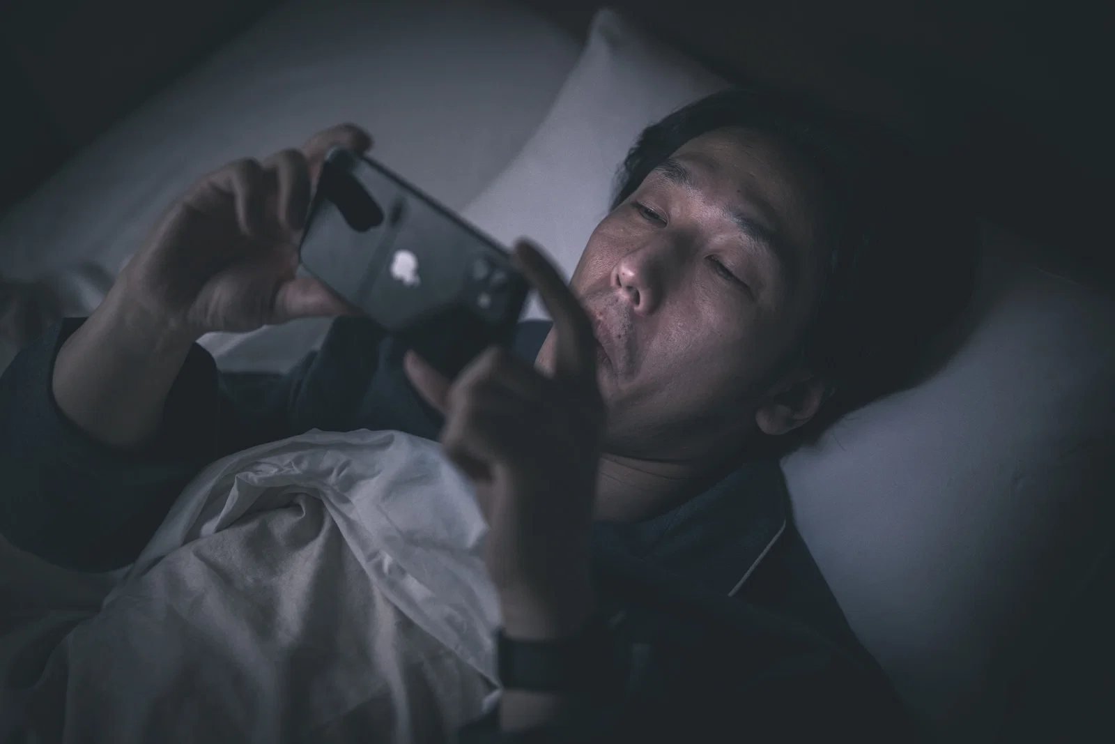 暗い室内でベッドに横たわりスマートフォンを操作するアジア系男性一人