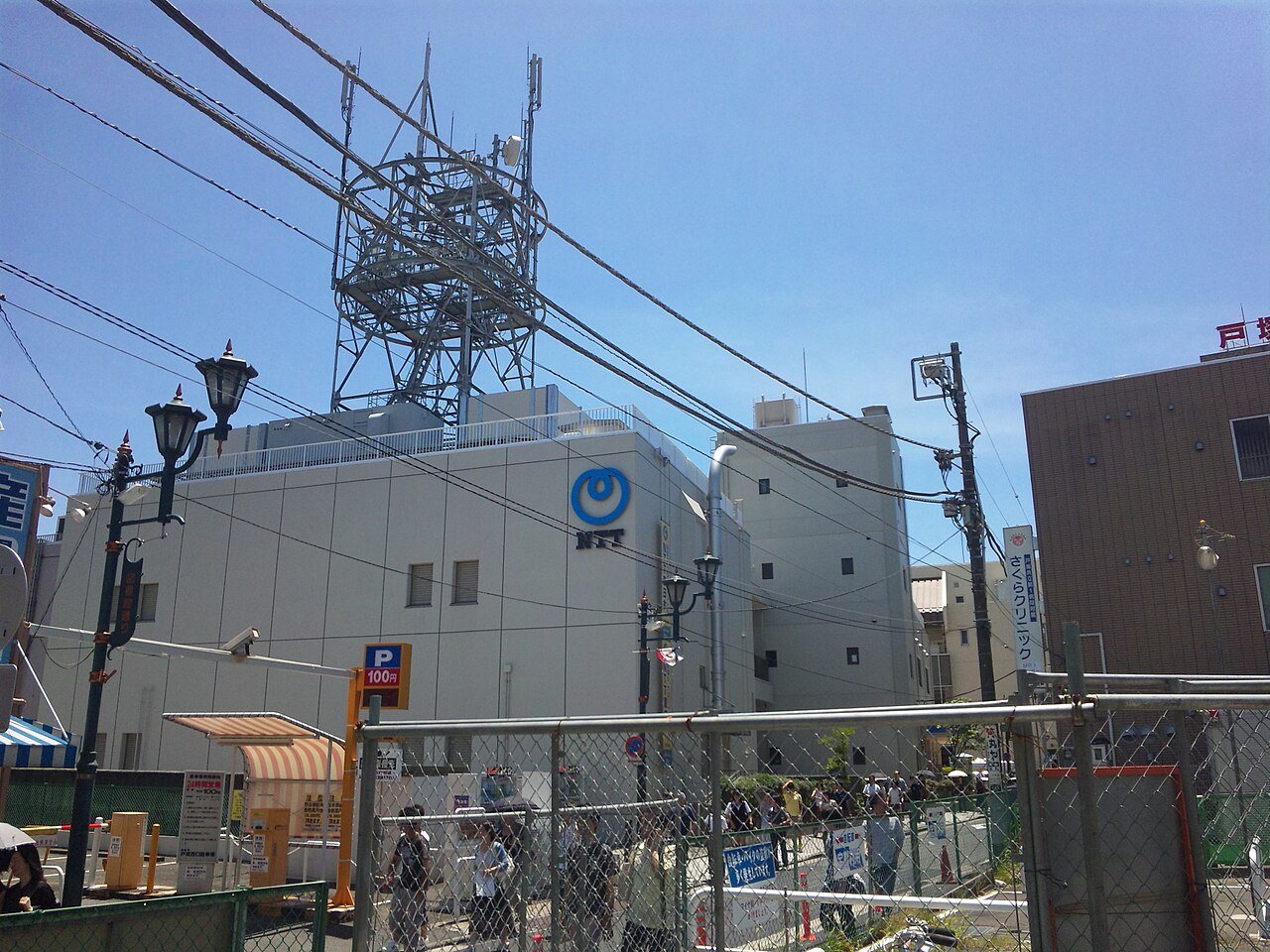 屋外、フェンス越しにNTTのロゴが付いた通信ビルと屋上鉄塔、通行人多数を写した写真